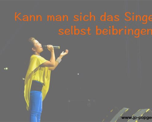 Kann man sich das Singen selbst beibringen Das Foto zeigt eine Sängerin in gelber Bluse und blauer Hose, die mit einem Mikrofon auf einer Bühne singt.