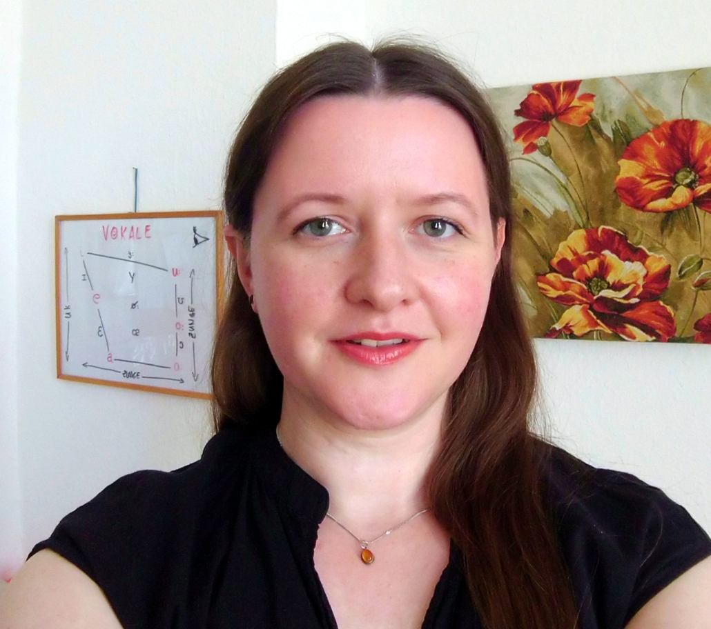 Ein Frau mit braunen Haaren und schwarzem Shirt vor einer Wand, an der Bilder von Mohnblumen und vom Vokaltrapez hängen.