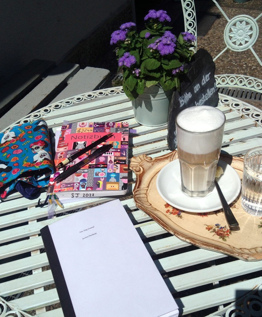 Ein Manuskript, Notizbuch, Stifte, lila Blumen im Top, Caffee Latte auf einem weißen Bistrotisch.