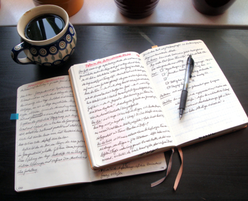 Auf einem dunklen Tisch liegen zwei aufgeschlagene und vollgeschriebene Notizbücher, darauf ein schwarzer Stift, daneben eine Tasse Kaffee.