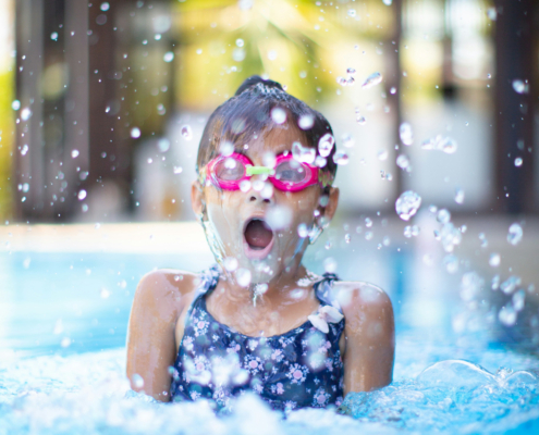 Ein Mädchen im blauen Badeanzug und mit pinker Taucherbrille taucht nach Luft schnappend aus dem blauen Wasser des Schwimmbads auf.
