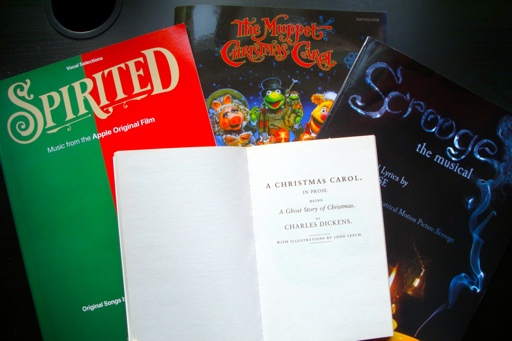 Das Deckblatt der Charles Dickens Weihnachtsgeschichte, A Christmas Carol, darunter drei Songbücher zu Adaptionen: v.l.n.r. Spirited, The Muppet Christmas Carol, und Scrooge: the musical, auf einer dunklen Tischoberfläche liegend.