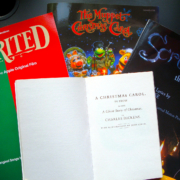 Das Deckblatt der Charles Dickens Weihnachtsgeschichte, A Christmas Carol, darunter drei Songbücher zu Adaptionen: v.l.n.r. Spirited, The Muppet Christmas Carol, und Scrooge: the musical, auf einer dunklen Tischoberfläche liegend.