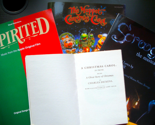 Das Deckblatt der Charles Dickens Weihnachtsgeschichte, A Christmas Carol, darunter drei Songbücher zu Adaptionen: v.l.n.r. Spirited, The Muppet Christmas Carol, und Scrooge: the musical, auf einer dunklen Tischoberfläche liegend.